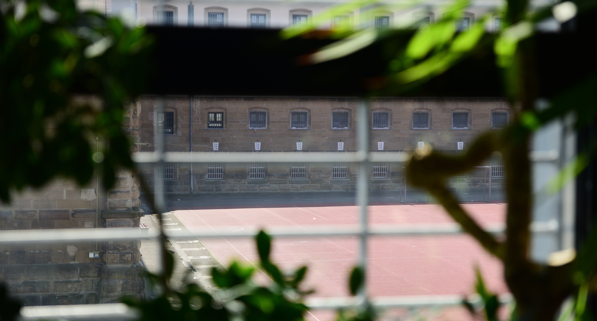 Blick durch einen Zaun auf ein Haftgebäude. Links und rechts befinden sich grüne Zweige.