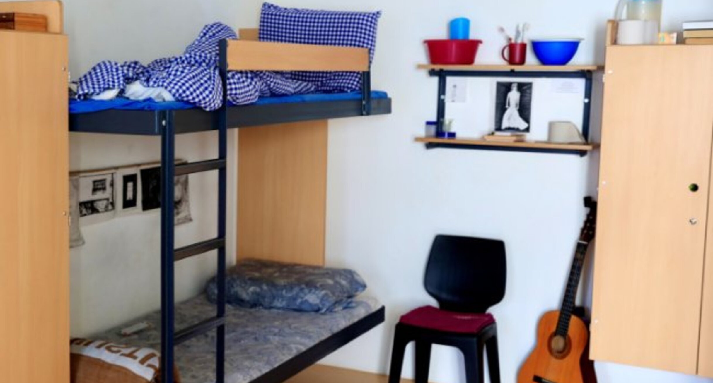 Zelle mit einem Doppelstockbett, Hängeregal, Stuhl und Schrank